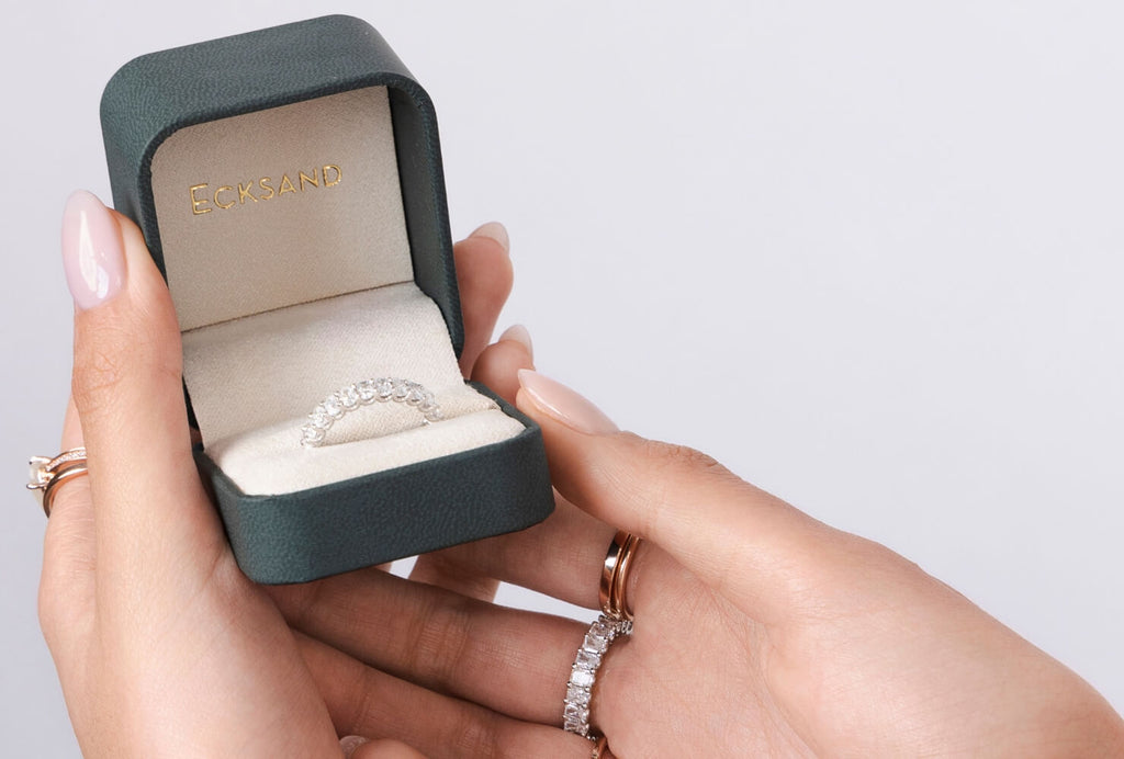 A woman holds an Ecksand box with a white gold diamond wedding ring, wearing a rose gold and a diamond eternity ring.