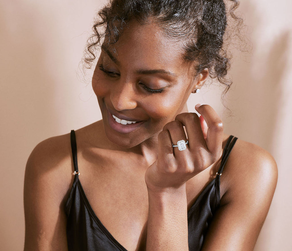 A woman wearing a Canadian-made emerald-cut diamond halo engagement ring and diamond stud earrings in 18K white gold.