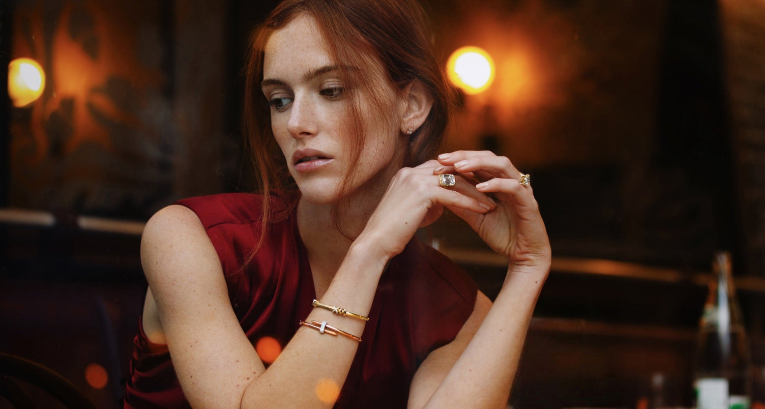 model wearing canadian-made ecksand 18k yellow gold emerald diamond engagement ring with 14k yellow and rose gold diamond bangle bracelets in a cafe in paris
