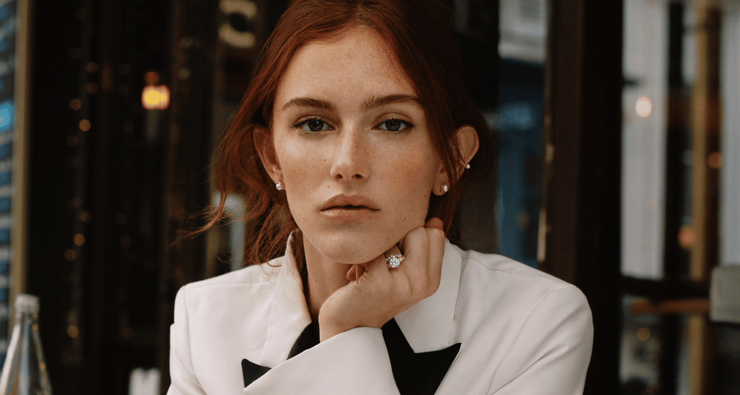 a model wearing a 18k rose gold diamond engagement ring with cluster setting and matching diamond wedding ring from ecksand's blossom collection in a white blazer with pearl earrings