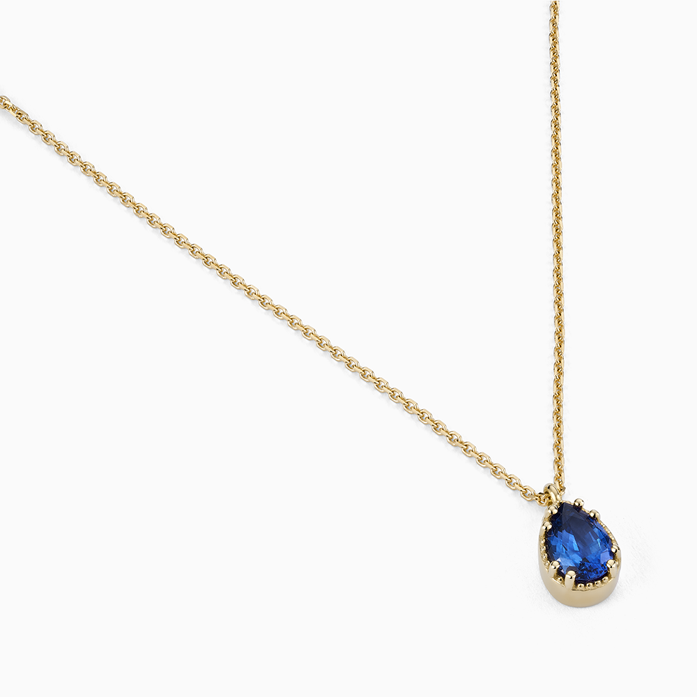 Angled view of Ecksand's Pear Blue Sapphire Stackable Necklace with Milgrain Halo in 18k Yellow Gold