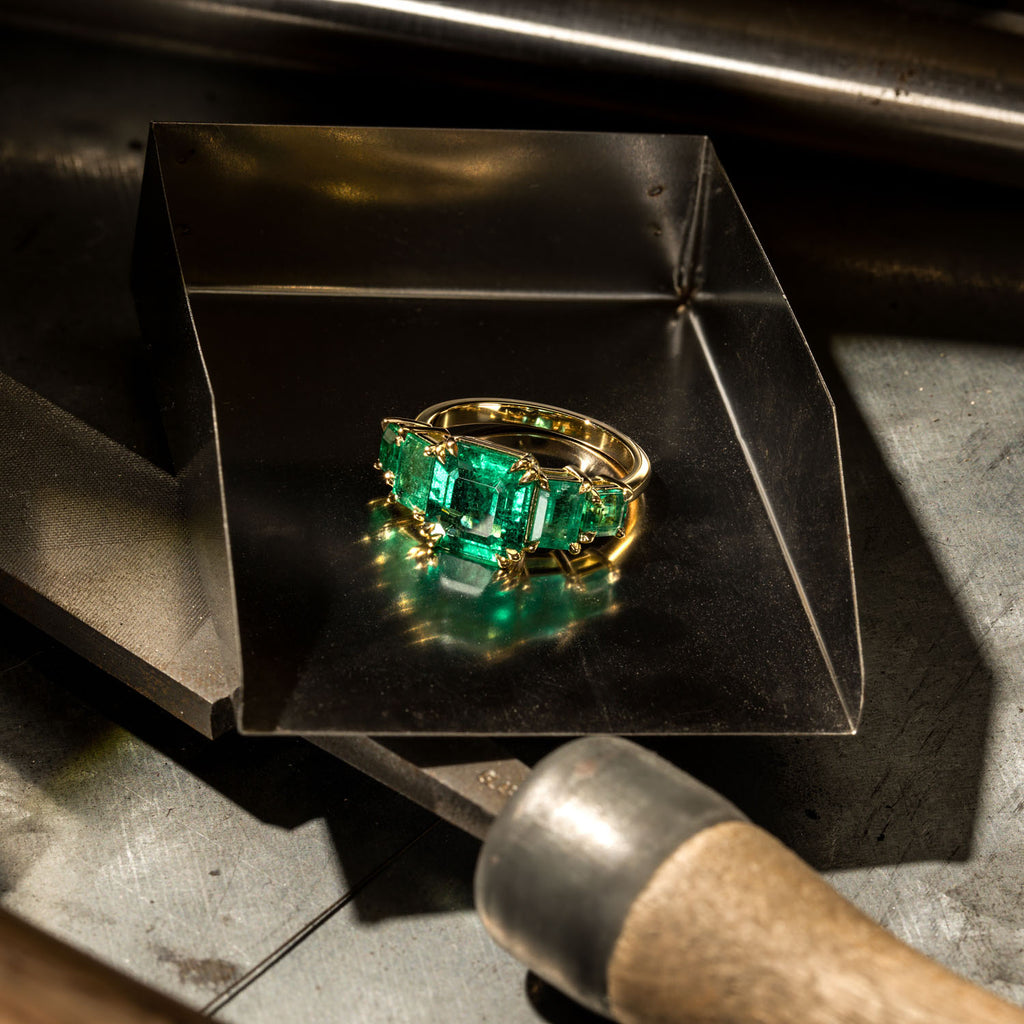 A Canadian-made three-stone emerald ring in 18K yellow gold sits in a jeweler's scoop on a workshop bench.