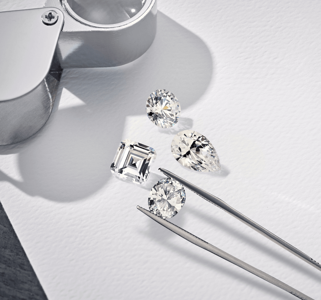 round, pear, and asscher cut diamonds in tweezers next to a loop in an ecksand workshop atelier on white background