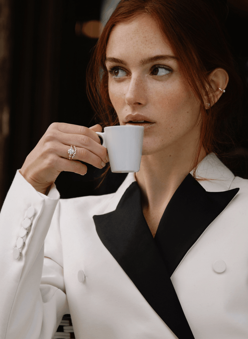 A woman with red hair wearing a white blazer with black lapels, holding a white coffee cup near her face. She is wearing a canadian-made 18k yellow gold blossom diamond engagement ring and curved diamond pave wedding ring on her finger.