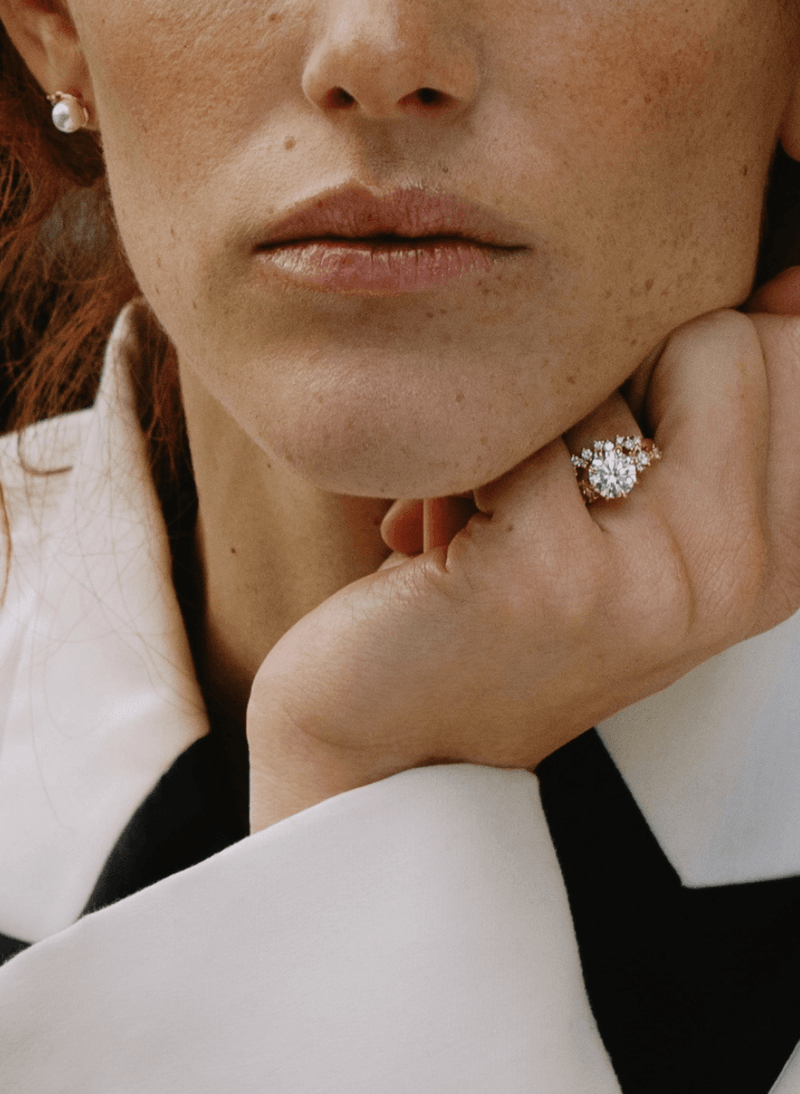 Close-up of a woman wearing a canadian-made 18k yellow gold round diamond engagement ring on her finger from ecksand's blossom collection, with a pearl stud earring visible on her ear. She is wearing a white and black collared shirt.
