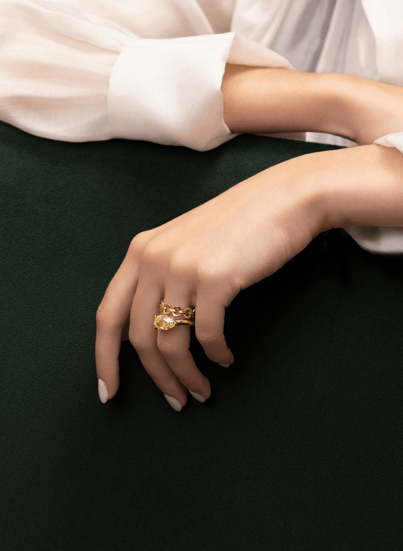A close-up of a person's hand wearing a canadian-made 18k yellow gold ring with a yellow pear cut diamond set east west on a curved band, resting on a dark green surface with a white sleeve in the background.