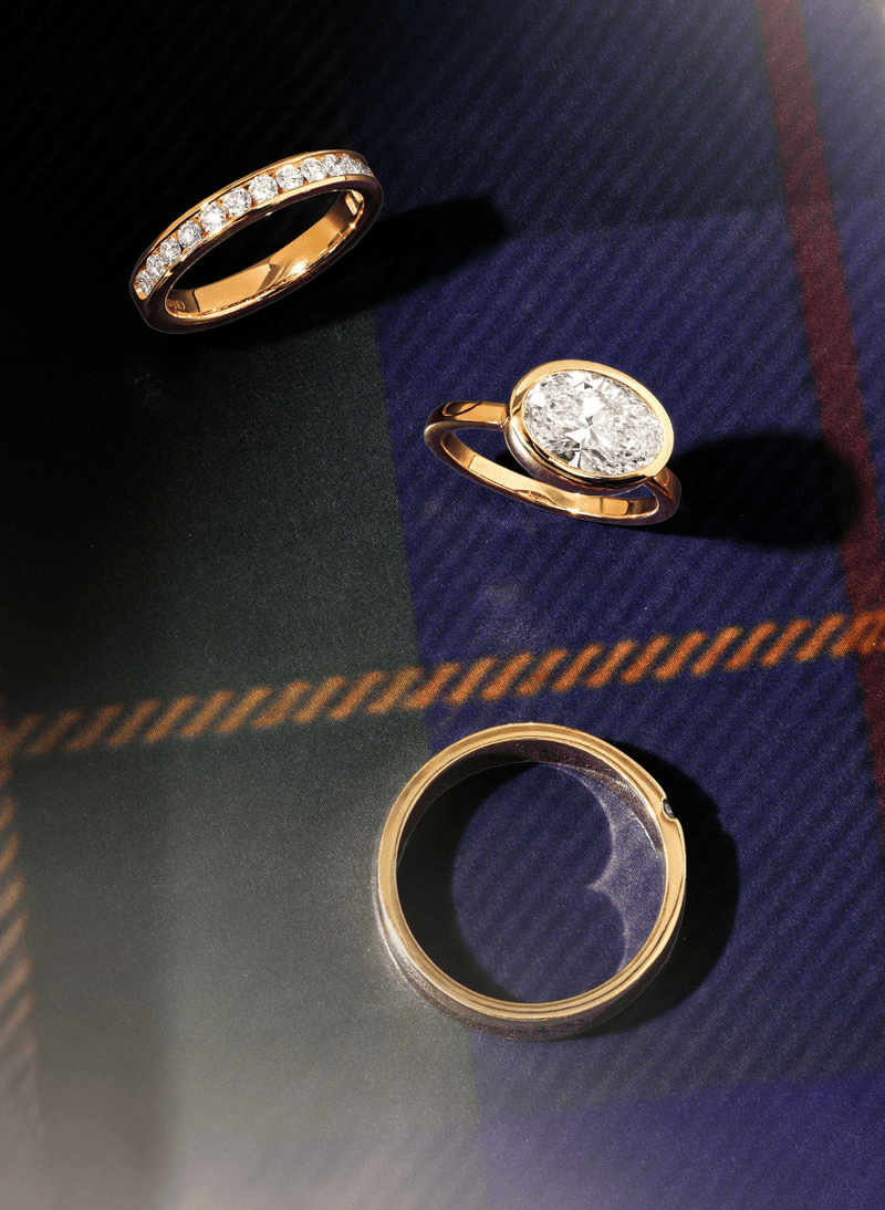 Three canadian-made 18k yellow gold rings displayed on a dark plaid fabric background: a pavé band ring, an oval east west bezel set diamond engagement ring, and a simple polished band ring.