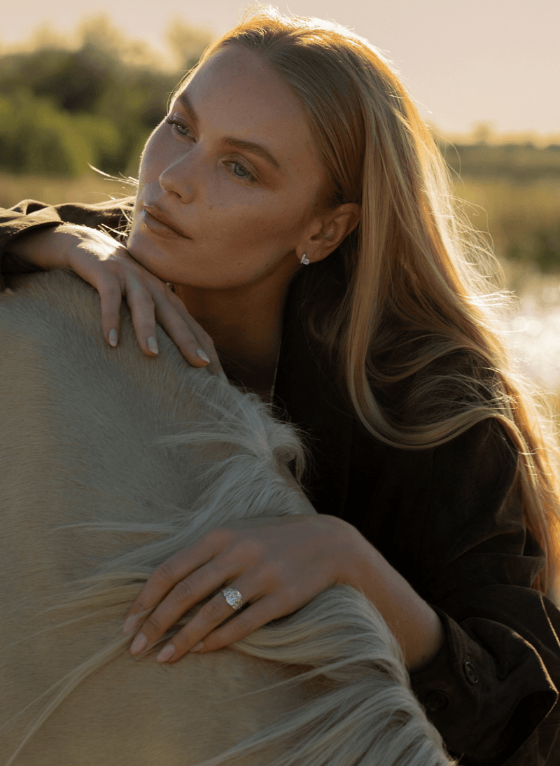 A woman with long blonde hair embraces a light-colored horse outdoors in warm golden hour light, wearing a canadian-made 18k white gold old mine diamond three stone engagement ring.