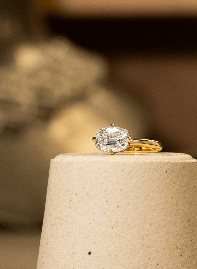 A canadian-made 18k yellow gold ring with a large old mine diamond in a east west twist prong setting, displayed on a textured beige cylindrical pedestal against a warm, blurred background.