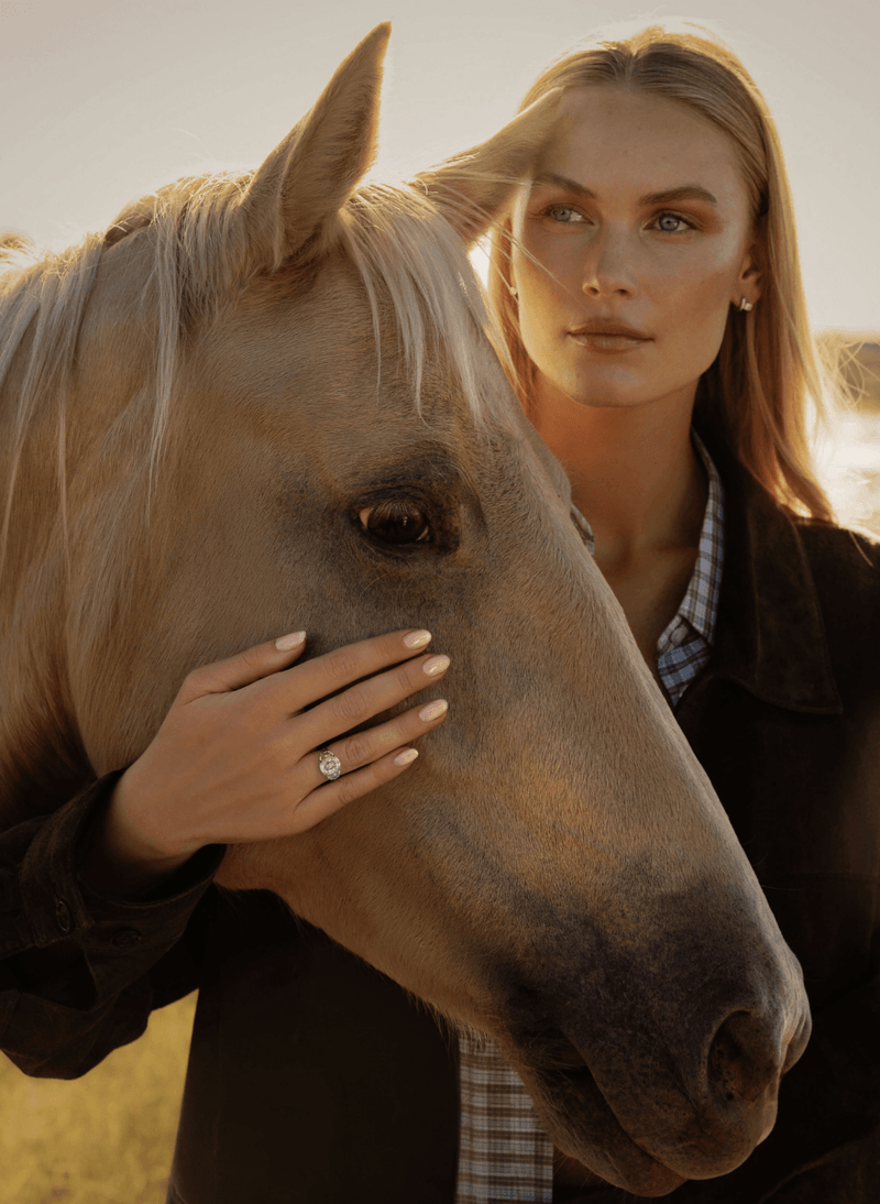 A woman with blonde hair stands behind a light brown horse, gently touching its face. She is wearing a dark brown jacket over a blue-and-white checkered shirt, and a canadian-made 18k white gold old mine diamond three stone engagement ring is visible on her finger.