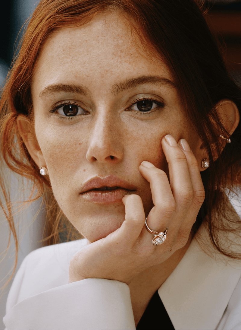 A close-up portrait of a woman with red hair resting her chin on her hand, wearing a white blazer, a canadian-made 18k white gold pear yellow diamond ring with a bezel setting, and small pearl stud earrings.
