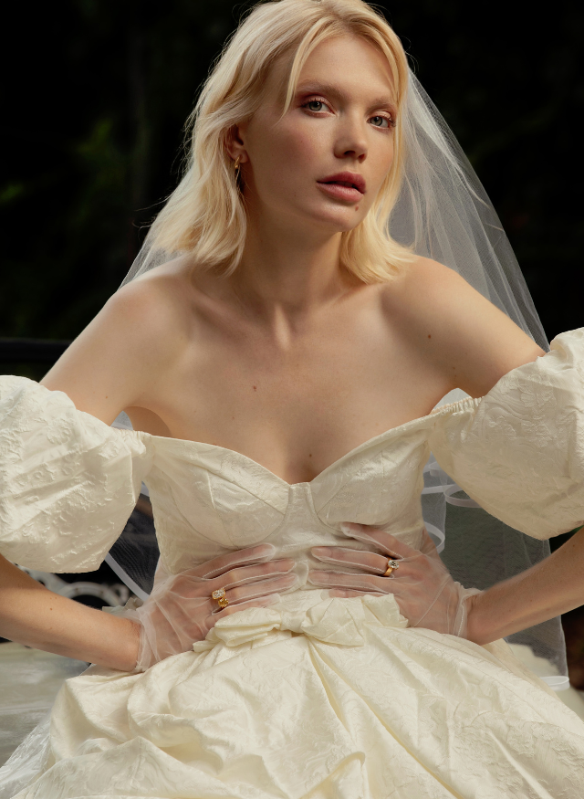 A blonde woman wearing a voluminous off-white wedding dress with puffed sleeves, sheer gloves, and a veil, posed against a dark outdoor background wearing canadian-made 18k yellow gold diamond engagement rings by ecksand.