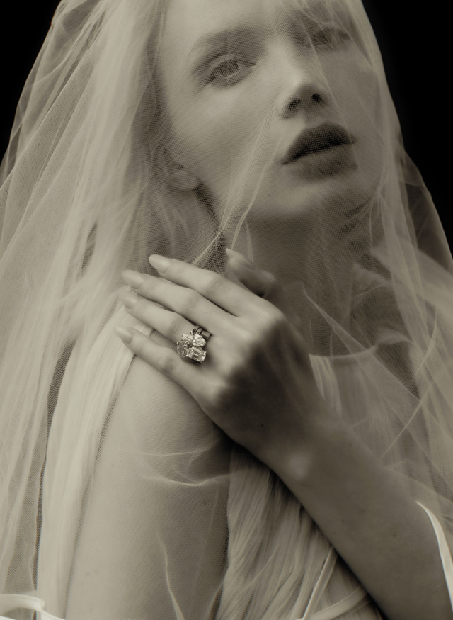 A close-up portrait of a woman wearing a sheer white veil and a three stacked canadian-made 18k yellow gold diamond engagement statement rings on one finger, shown in a monochrome sepia-toned photograph against a dark background.