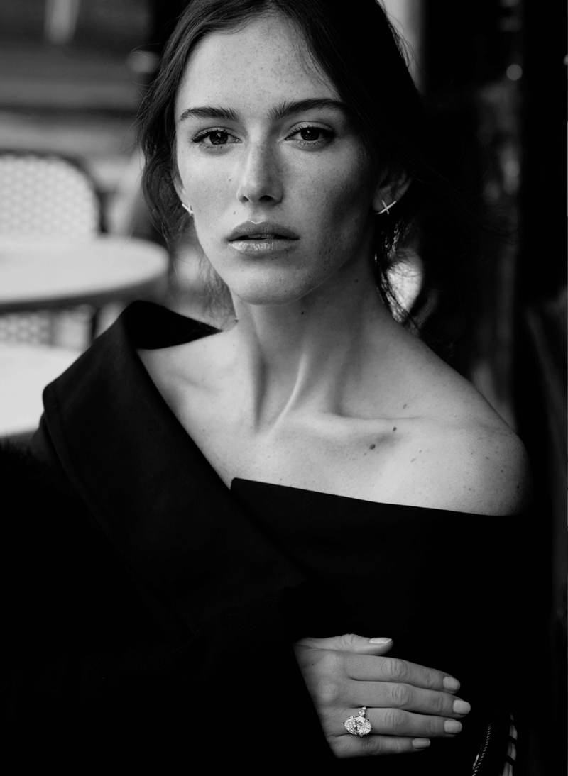 A black and white portrait of a woman wearing a black off-the-shoulder top and a canadian-made 18k white gold oval diamond engagement ring set against a softly blurred background.
