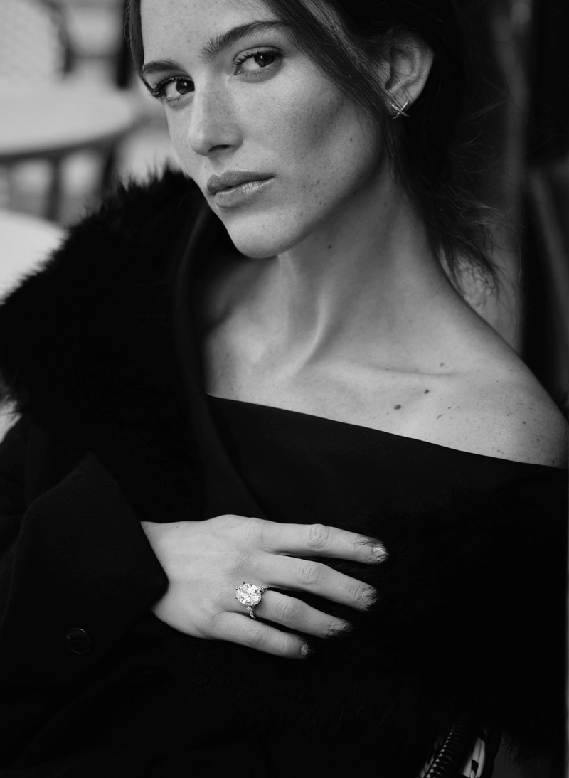 A black and white portrait of a woman wearing a black off-the-shoulder top and a canadian-made 18k white gold oval diamond engagement ring set against a softly blurred background.