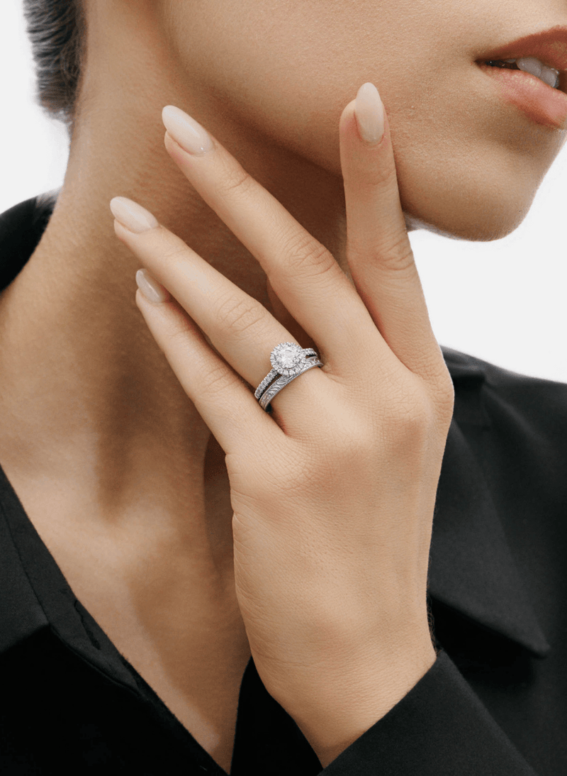 A close-up lifestyle shot of a model wearing a canadian-made 18k white gold engagement ring with a round-cut center stone in a halo setting and a matching pavé band, set against a clean white background.