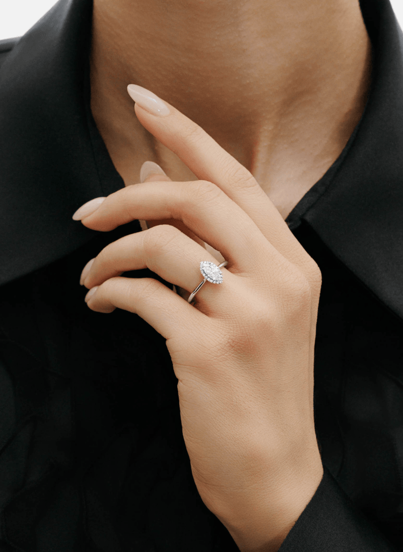 A close-up of a person's hand wearing a canadian-made 18k white gold ring with a marquise cut diamond in a halo setting, set against a black collared shirt.