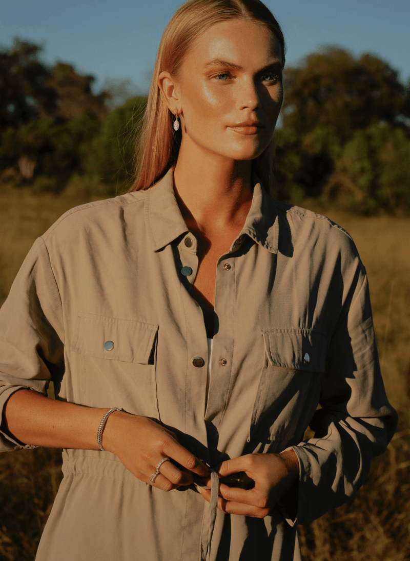 A woman outdoors in warm golden-hour light wearing a light beige button-up shirt dress with chest pockets and a drawstring waist wearing 18k white gold diamond fine jewelry from ecksand's arctic dragon collection.
