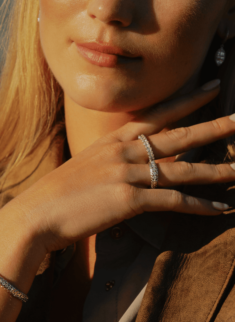 A close-up lifestyle shot of a woman wearing canadian-made 18k white gold diamond band rings on her fingers, with warm sunlight highlighting her face, hand, and brown jacket.