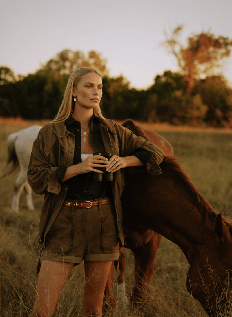 A woman stands in a grassy field at sunset wearing an olive green utility jacket and matching shorts over a black shirt with canadian-made 18k white gold reversible diamond jewelry from ecksand's aura collection, with a brown horse beside her and a white horse in the background.