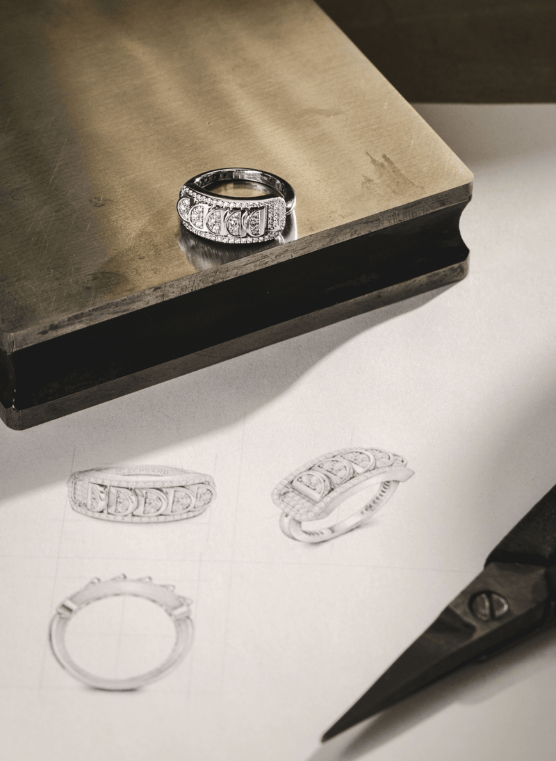 An 18k white gold reversible diamond ring with an intricate openwork filigree pattern sits on a metallic brass block, with hand-drawn sketches of the same ring design on white paper below and a pair of scissors in the foreground.