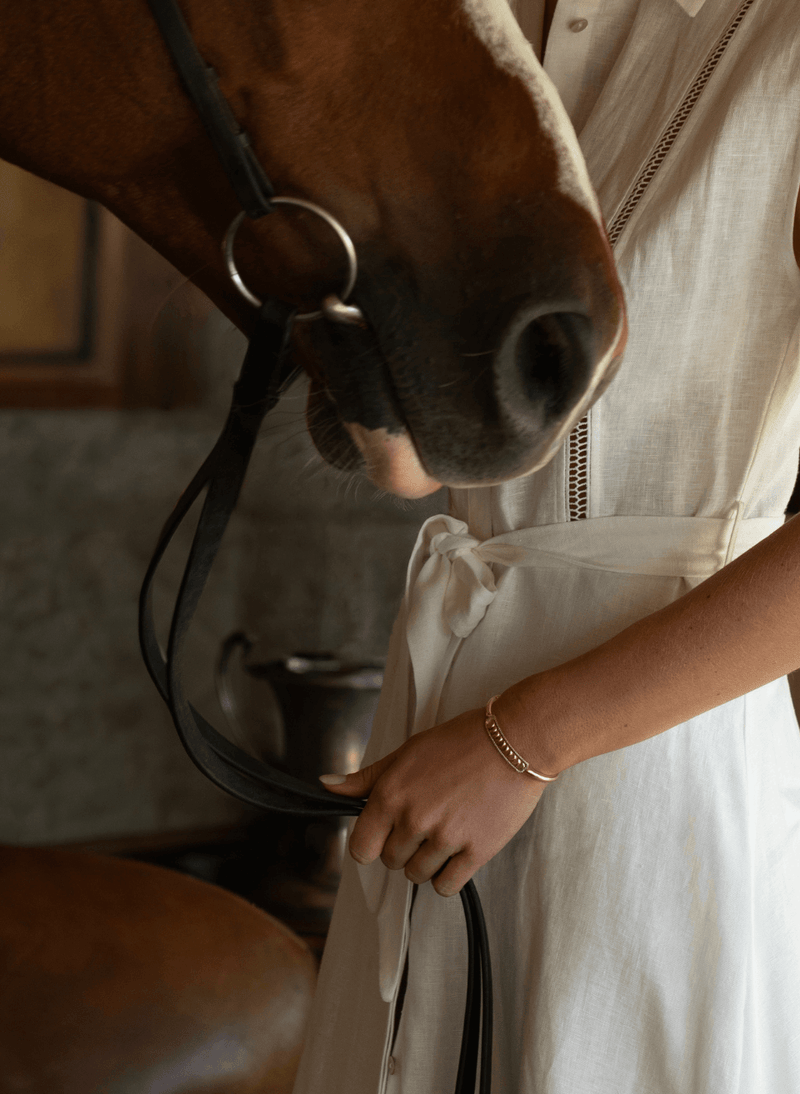 A close-up lifestyle shot of a person in a white linen dress holding the reins of a brown horse with a bridle. The person is wearing a delicate 18k yellow gold reversible diamond bracelet, and the background is a softly blurred rustic indoor setting.
