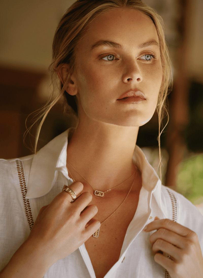 A close-up lifestyle shot of a woman wearing delicate canadian-made 18k yellow gold reversible necklaces and rings, featuring a small rectangular gold pendant on one chain and a gold ring on her finger, set against a warm, softly blurred background.