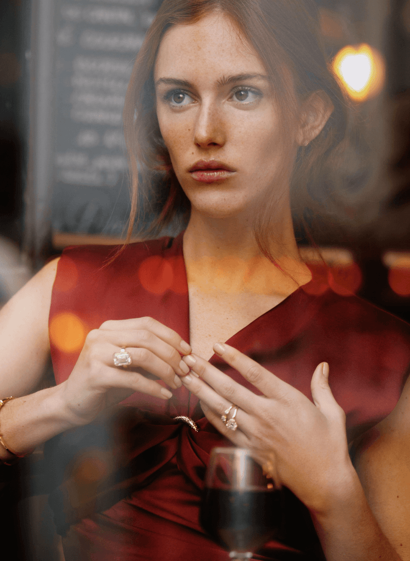 A woman in a sleeveless deep red dress sits behind a window, looking off-camera with a glass of red wine in the foreground and warm bokeh lights in the background. she is wearing canadian-made 18k yellow gold diamond rings.