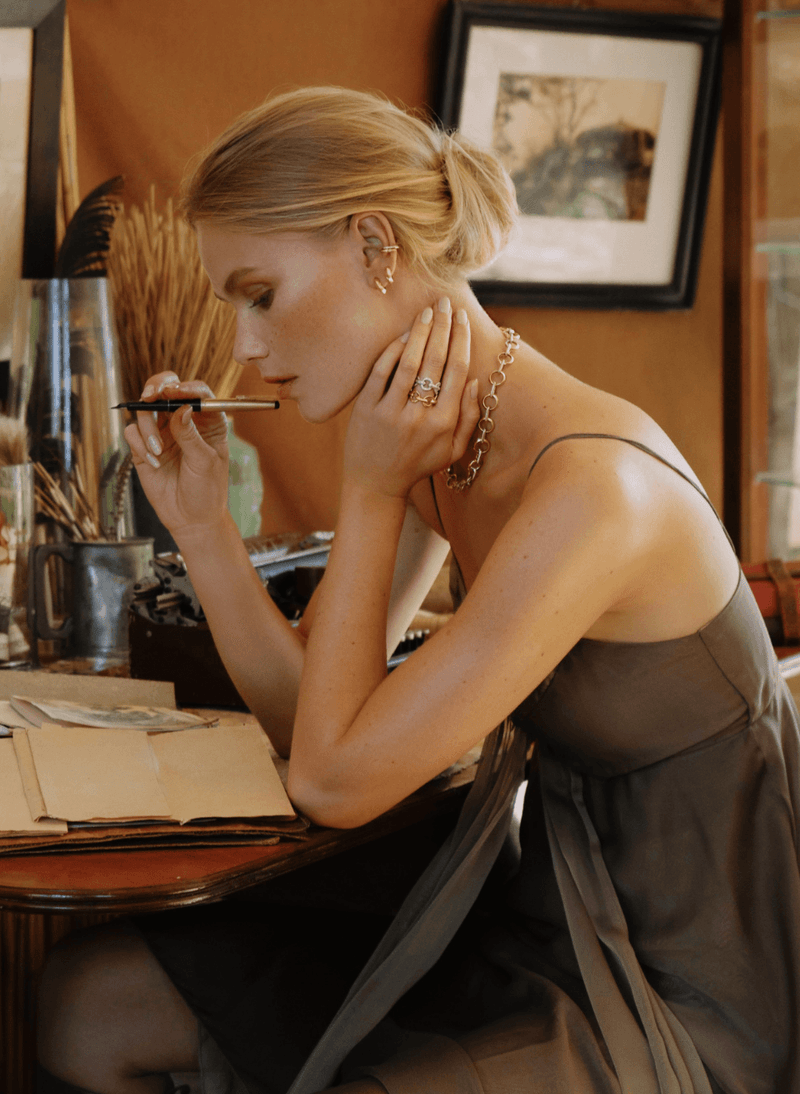 A blonde woman in a dark gray dress sits at a wooden table, holding a pen to her chin in a warm, vintage-styled room. She is wearing a canadian-made 18k yellow gold chain necklace and gold rings from ecksand's duel chain collection.