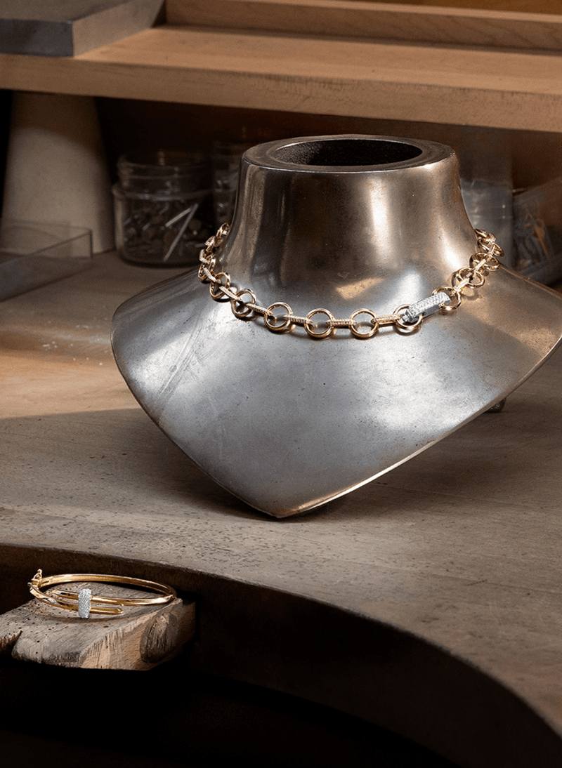 A canadian-made 18k gold chain necklace on a brushed silver metal bust, with a matching 18k yellow gold bangle bracelet resting on a wooden surface in the foreground from ecksand's duel collection.