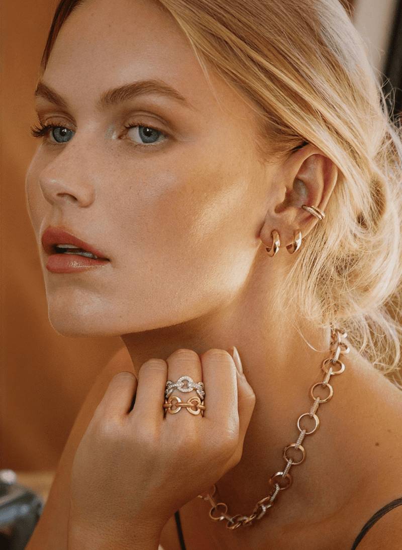 Close-up of a blonde woman wearing multiple canadian-made 18k yellow and white gold jewelry pieces, including hoop earrings, a chain necklace, and stacked rings on her hand from ecksand's duel chain collection against a warm brown background.