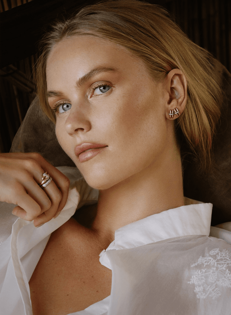 A close-up portrait of a woman wearing a canadian-made 18k yellow gold small gold stud earring with a diamonds and a 18k white gold wrap diamond ring on her finger, dressed in a white collared blouse against a dark, warm-toned background.