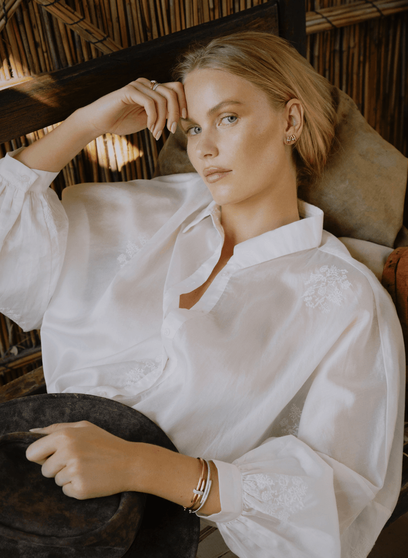 A woman with blonde hair wearing a white long-sleeve embroidered blouse, posing in a rustic setting with bamboo walls wearing canadian-made 18k yellow gold fine jewelry from ecksand's duel collection.