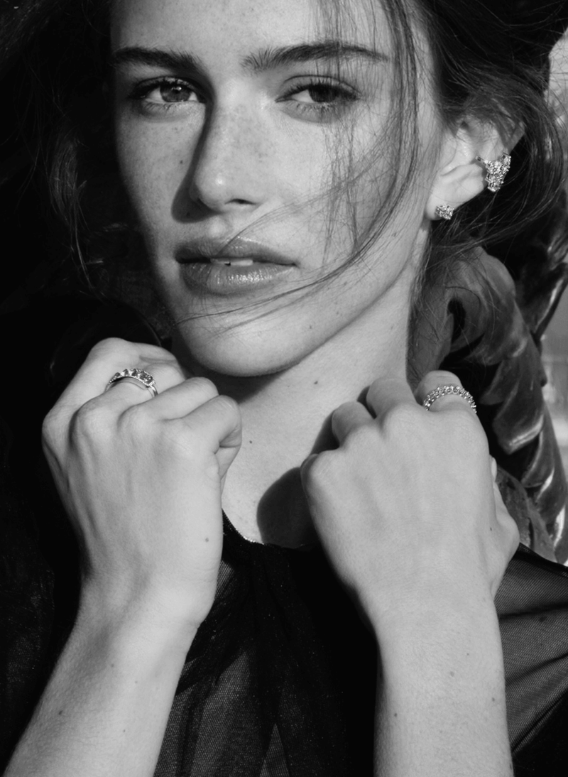 A black and white close-up portrait of a woman wearing multiple canadian-made 18k white gold diamond rings on her fingers and a diamond conch earring earring, with her hands near her neck.