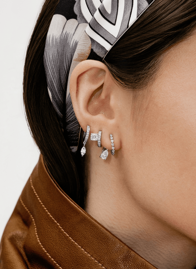 A close-up of a model's ear wearing a stack of canadian-made 18k white gold diamond earrings including asscher diamond hoops, a marquise drop hoop, and a diamond pave hoop stud jacket earrings. The model has dark brown hair styled with a black, white, and gray patterned silk headband and is wearing a brown leather jacket.