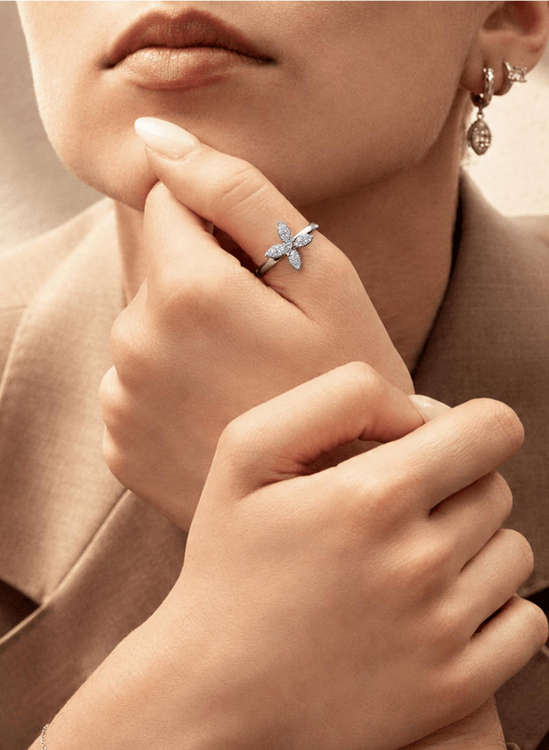 A close-up lifestyle shot of a model wearing a delicate canadian-made 18k white gold diamond ring with a butterfly design on the thumb. The model is wearing a beige blazer and matching earrings, with warm, soft lighting highlighting the jewelry.