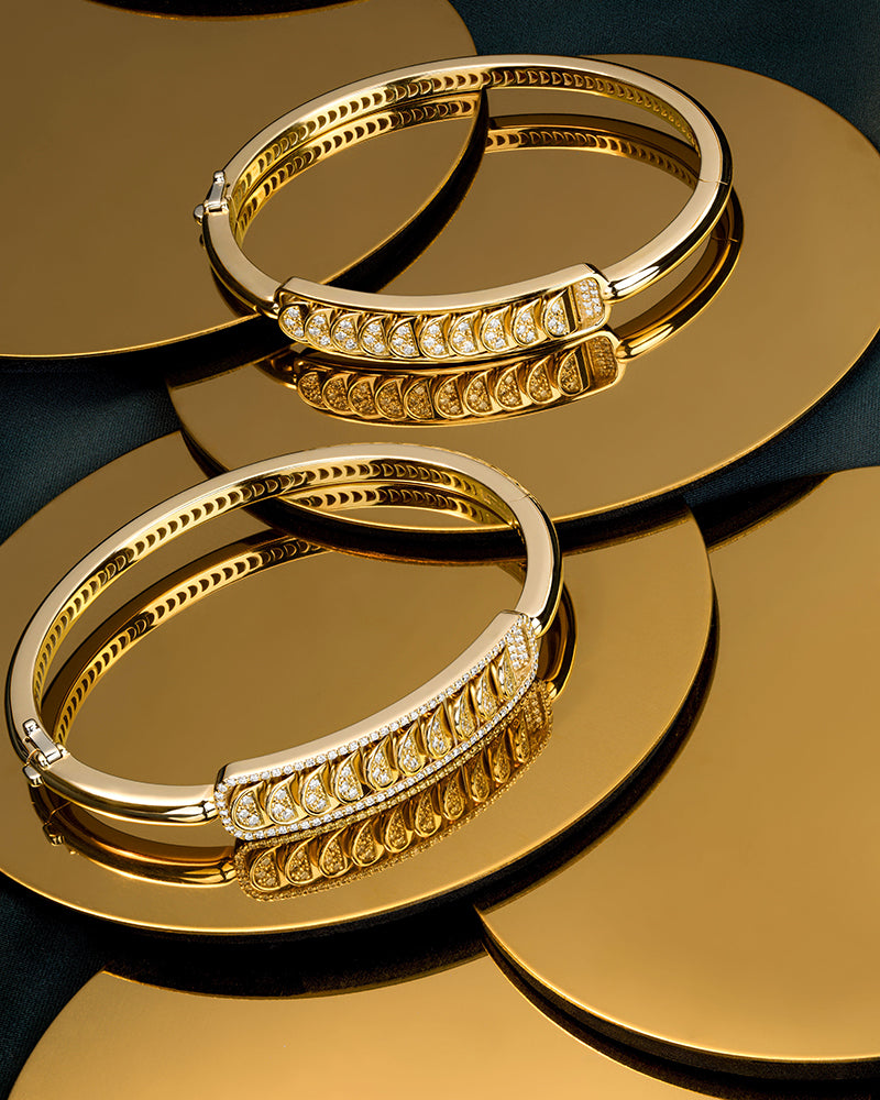 Two diamond bangle bracelets in 18k yellow gold elegantly positioned on a reflective gold surface.