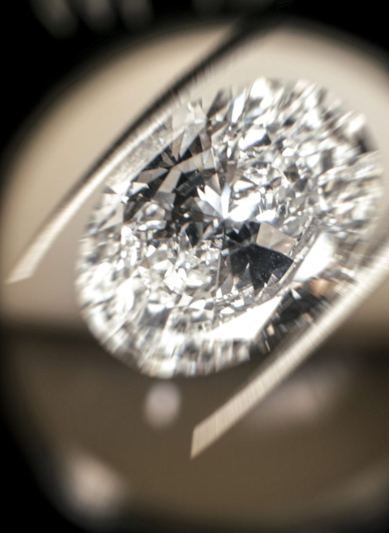 A close-up macro shot of a brilliant oval-cut diamond held in a metal tweezer against a dark, blurred background in a canadian atelier.