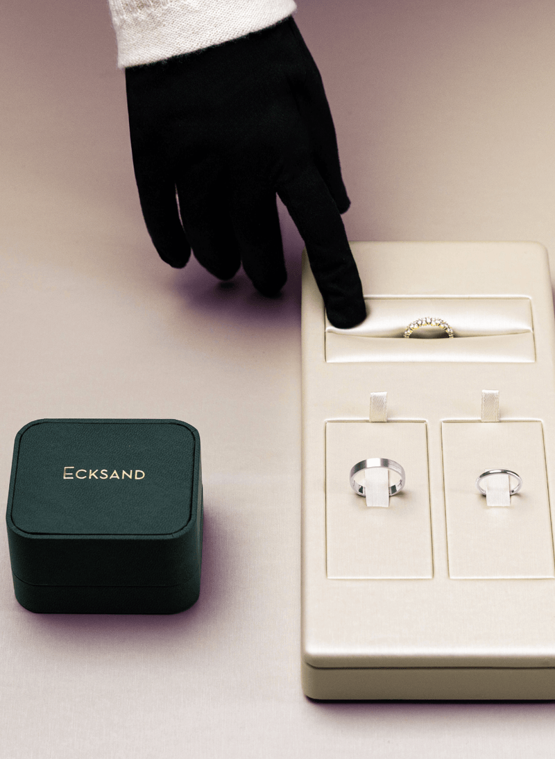 A hand wearing a black glove points toward a cream-colored ring display box containing three canadian-made rings, with an open dark green Ecksand jewelry box beside it on a soft beige background.