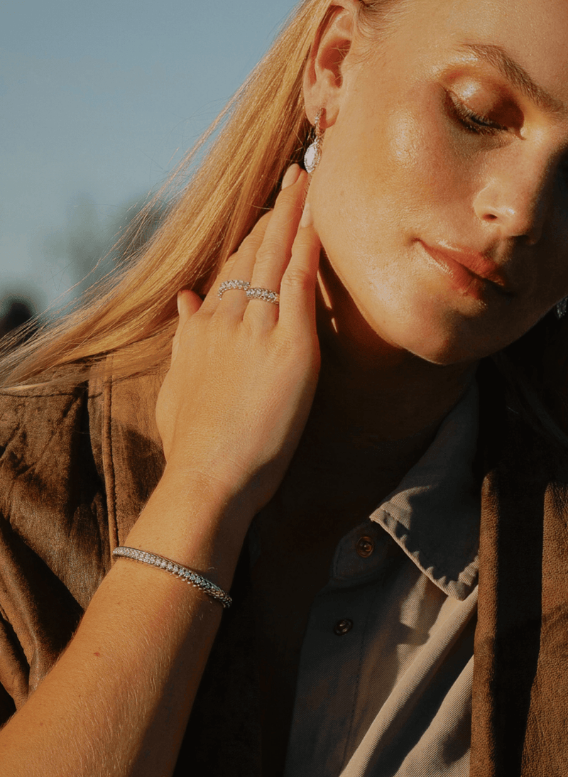 A close-up lifestyle shot of a woman with blonde hair wearing a bangle bracelet on her wrist, a matching canadian-made 18k white gold eternity ring on her finger, and a diamond hoop earring, set against a warm golden-hour outdoor background.