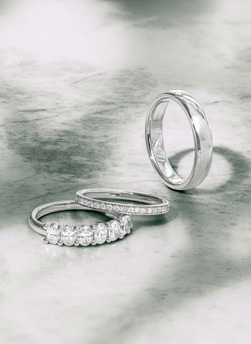 Three canadian-made 18k white gold rings displayed on a textured gray surface: a plain band ring standing upright, a diamond-accented band ring lying horizontally, and another diamond-accented band ring lying diagonally.