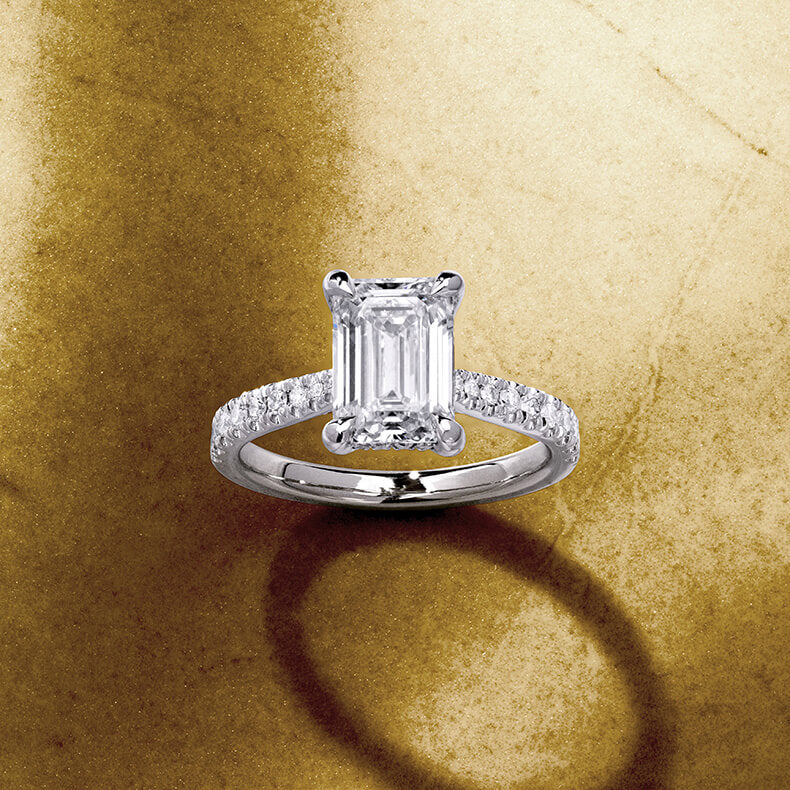 Canadian-made emerald-cut diamond engagement ring in 18K white gold with diamond pavé band on a textured background